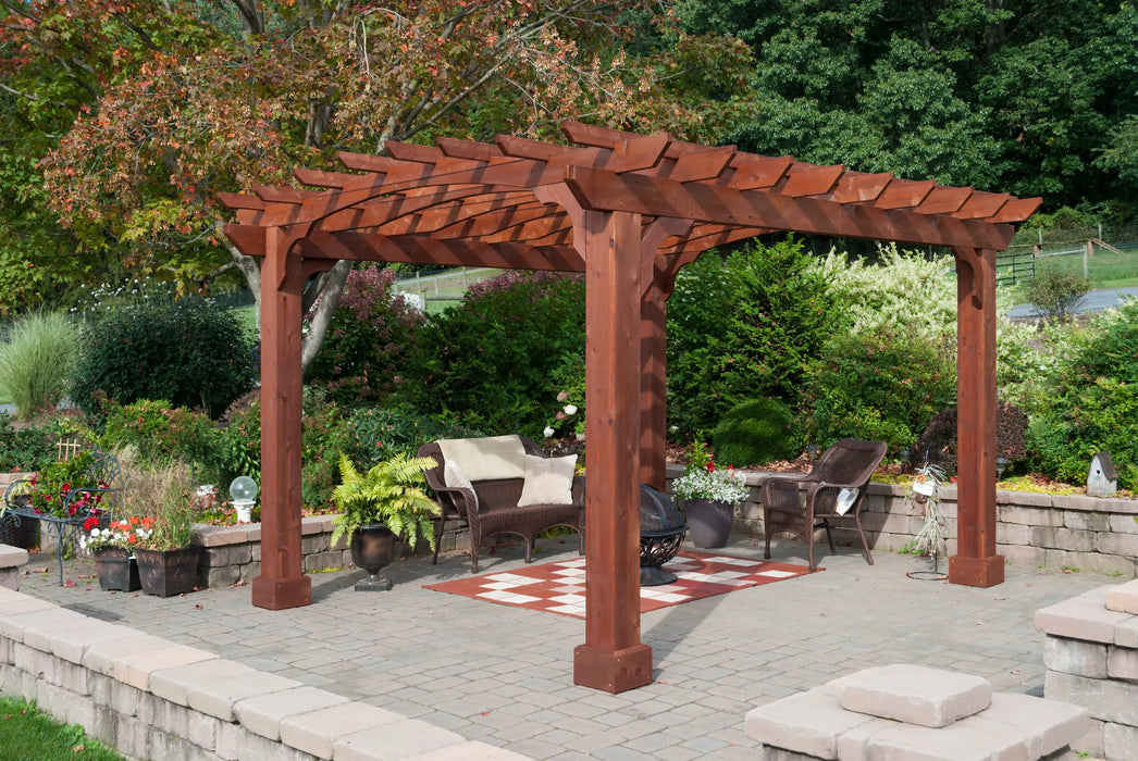A dark-stained wooden pergola stands on a patio made of gray pavers, with two wicker chairs and a fire pit visible beneath it. Lush greenery surrounds the patio.