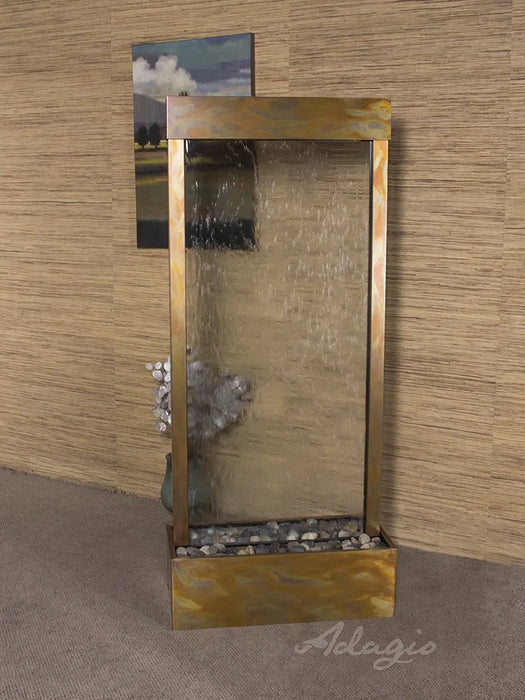 A free standing water fountain, a beautiful adagio water feature. The "Harmony River," features a wide, textured glass panel where water gently cascades down into a shallow basin filled with smooth, grey river stones.