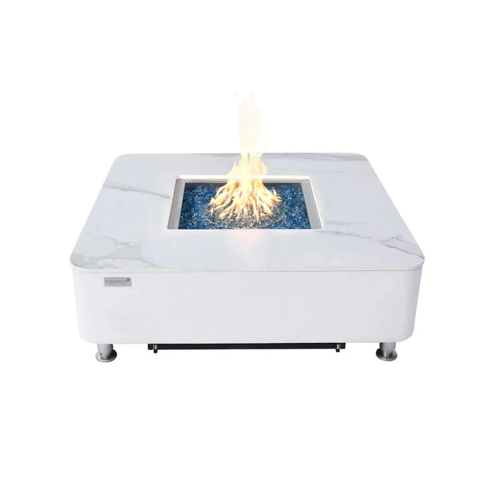 Modern white square fire pit table with a marble-look top and a stainless steel burner, lit with a flickering flame. The table has a white base and is elevated on small silver feet.