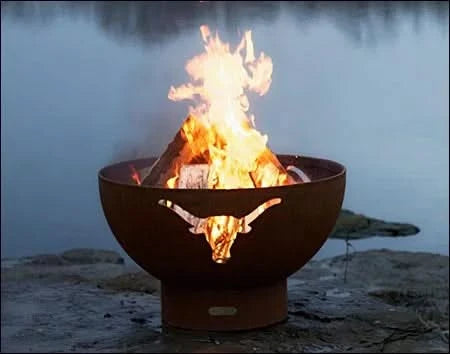 A wood-burning fire pit with a longhorn cutout design, placed on a rocky shoreline near a lake with a misty background. The fire is burning brightly.