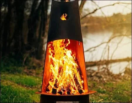A tall, conical wood-burning fire pit with flames burning inside. The fire pit is surrounded by green grass and is near a body of water. There is a deer logo on the side of the fire pit.