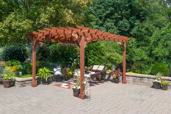 A brown wooden pergola with a slatted top sits on a stone patio. Two chairs and a small table are placed underneath it.