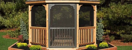 Close-up of a cedar gazebo with a black gazebo screen door and wooden railing, set in a garden with mulch and plants.