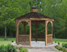 A wooden octagonal gazebo with a shingled roof, decorative cupola, and lattice detailing on the walls. The gazebo is surrounded by lush greenery and a paved patio.