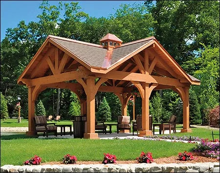 A rustic wooden ramada structure with a gabled roof and a copper cupola. The gazebo is surrounded by green grass and colorful flowers. There are chairs and a table inside the gazebo.