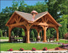 A rustic wooden ramada structure with a gabled roof and a copper cupola. The gazebo is surrounded by green grass and colorful flowers. There are chairs and a table inside the gazebo.