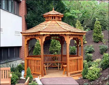 A wooden octagonal gazebo with a shingled roof and a decorative cupola. The gazebo features latticework detailing on the walls and a spacious interior with a built-in bench.