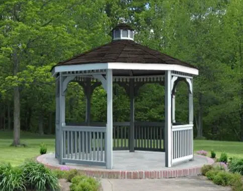 A gray octagonal vinyl gazebo with a classic design stands on a concrete pad surrounded by lush greenery. The gazebo features a shingled roof with a decorative cupola, white latticework railings, and built-in benches.