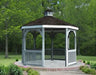 A gray octagonal vinyl gazebo with a classic design stands on a concrete pad surrounded by lush greenery. The gazebo features a shingled roof with a decorative cupola, white latticework railings, and built-in benches.