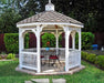 A white octagonal gazebo with a classic design stands on a brick patio surrounded by greenery. The gazebo features a gray shingled roof with a decorative cupola, white latticework railings, and built-in benches. A small table with chairs sits in the center of the gazebo. Trees and shrubs frame the gazebo in the background.