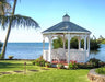 A white octagonal gazebo with a classic design stands on a green lawn overlooking a calm blue ocean. The gazebo features decorative railings, a shingled roof with a cupola, and built-in benches. A palm tree and lush greenery surround the gazebo.