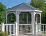 A white octagonal vinyl gazebo with a classic design stands on a deck overlooking a lush green lawn. The gazebo features a gray shingled roof, white latticework railings, and screened-in sides with a door. Flowers and shrubs surround the gazebo.