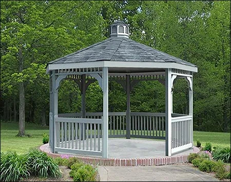 A gray octagonal gazebo with a classic design stands on a brick patio surrounded by greenery. The gazebo features a shingled roof with a decorative cupola, white latticework railings, and built-in benches. Trees and shrubs frame the gazebo in the background.