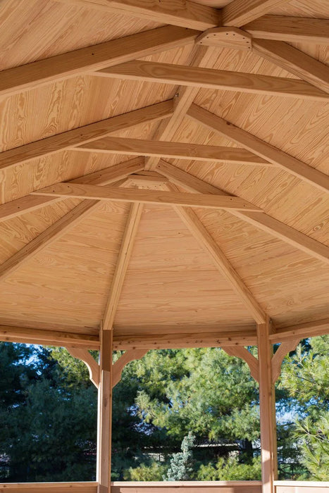 A person enjoying a meal at a wooden table inside a large, octagonal gazebo. The gazebo has a shingled roof and open sides, with greenery visible in the background.