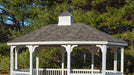 White vinyl octagon gazebo with a sloped roof, featuring decorative roof trim and exposed wooden rafters. The gazebo is surrounded by lush green trees.