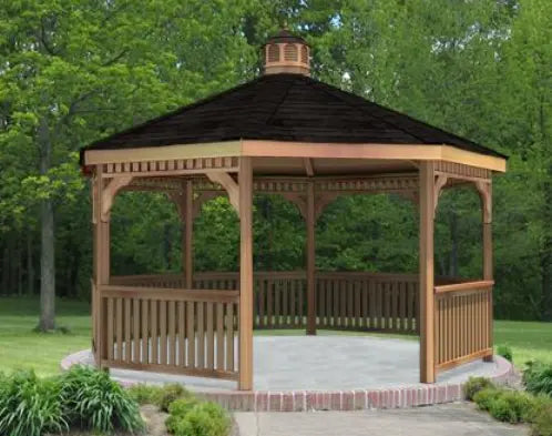 A large octagonal wooden gazebo with a shingled roof and a decorative cupola. The gazebo features a spacious interior with open latticework walls and is surrounded by a brick patio and lush greenery.