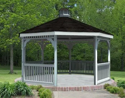 A white octagonal vinyl gazebo with a classic design stands on a brick patio surrounded by greenery. The gazebo features a black shingled roof with a decorative cupola, white latticework railings, and built-in benches. Trees and shrubs frame the gazebo in the background.