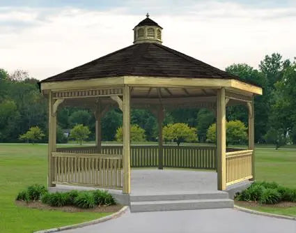 A large octagonal gazebo with a classic design. It features a shingled roof with a decorative cupola, and intricate latticework railings surrounding the open structure. The gazebo is painted a light brown color and sits on a concrete pad surrounded by greenery.