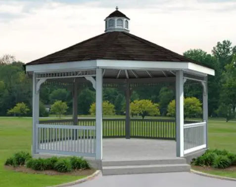 A white octagonal gazebo with a classic design stands on a concrete pad surrounded by lush greenery. The gazebo features a black shingled roof with a decorative cupola, white latticework railings, and built-in benches. Trees and shrubs frame the gazebo in the background.