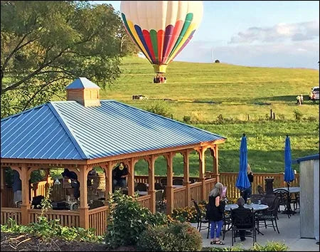 A wooden gazebo with a blue roof sits on a grassy area with people gathered around tables. In the background, a colorful hot air balloon floats over rolling hills