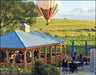 A wooden gazebo with a blue roof sits on a grassy area with people gathered around tables. In the background, a colorful hot air balloon floats over rolling hills