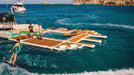 People on a floating platform in the ocean with boats and cliffs in the background