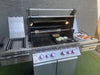 A Napoleon gas grill with the hood open. The reversible griddle is cooking eggs and bacon, with a plate of cooked bacon on the side.