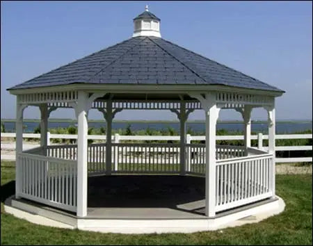 A white hexagonal gazebo with a gray shingled roof stands on a raised platform. The gazebo features decorative white railings and offers a view of a body of water.