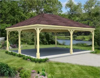 large pine wood gazebo with a gabled roof and a concrete base. The gazebo is surrounded by lush greenery and a lake is visible in the background.