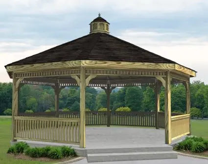 A large dodecagon gazebo with a classic design. It features a shingled roof with a decorative cupola, and intricate latticework railings surrounding the open structure. The gazebo is painted a light brown color and sits on a concrete pad surrounded by greenery.