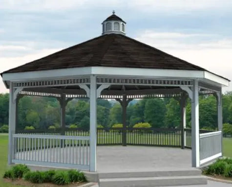 A white octagonal vinyl gazebo with a classic design. It features a shingled roof with a decorative cupola, white latticework railings, and built-in benches. Trees and shrubs frame the gazebo in the background.