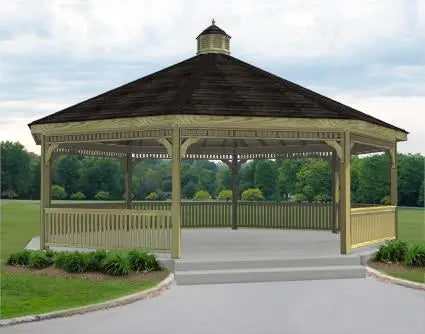 A large dodecagon gazebo with a classic design. It features a shingled roof with a decorative cupola, and intricate latticework railings surrounding the open structure. The gazebo is painted a light brown color, and it has a raised floor with steps leading up to it. The gazebo is surrounded by a concrete pad and lush greenery.