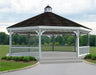 A large white octagonal gazebo with a classic design stands on a paved walkway surrounded by lush greenery. The gazebo features a black shingled roof with a decorative cupola, white latticework railings, and built-in benches. Trees and shrubs frame the gazebo in the background.