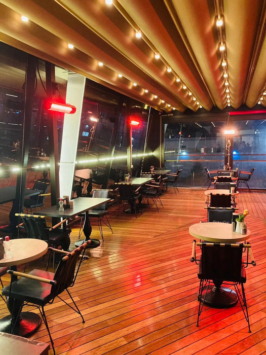 A well-lit restaurant patio with tables and chairs arranged along a wooden deck. Electric patio heaters provide warmth, and string lights add a festive ambiance.