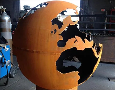 A spherical wood-burning fire pit shaped like the Earth. The continents are cut out of the metal, creating a unique and eye-catching design. The fire pit is a rusty orange color and is placed on a metal stand.