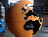 A spherical wood-burning fire pit shaped like the Earth. The continents are cut out of the metal, creating a unique and eye-catching design. The fire pit is a rusty orange color and is placed on a metal stand.