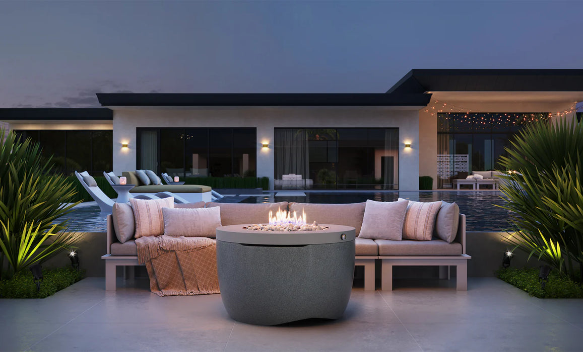 Gray Ledge Lounger Halo Fire Pit on a modern patio with a beige sectional sofa, pool, and contemporary house in the background at dusk.