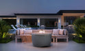 Gray Ledge Lounger Halo Fire Pit on a modern patio with a beige sectional sofa, pool, and contemporary house in the background at dusk.