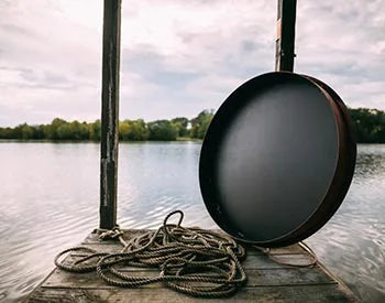 A round, black fire pit lid leaning against a wooden post on a dock. There is a pile of rope next to it.