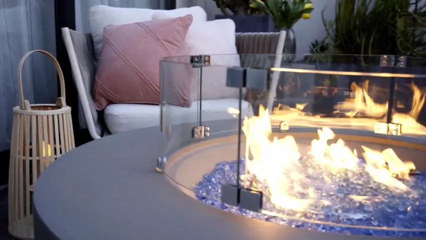 Close-up of a modern fire table with a protective glass screen, flames burning over blue glass pieces, with seating visible behind.