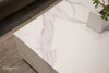 A close-up showing the corner of a rectangular coffee table with a white marble porcelain top featuring light grey veining. A white square pot with a small green plant sits on the tabletop.