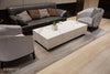 A rectangular coffee table with a white marble porcelain top featuring grey veining, and a matching white base, sits in a living room. A potted plant and two white cups are placed on the table.