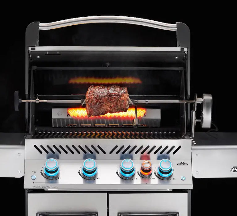 A close-up image of a stainless steel gas grill with a rotisserie kit installed. A large cut of meat is rotating on the spit rod, cooking over a bed of glowing infrared burners. The grill has a stainless steel hood with a handle, and the control knobs are visible at the bottom.