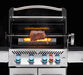 A close-up image of a stainless steel gas grill with a rotisserie kit installed. A large cut of meat is rotating on the spit rod, cooking over a bed of glowing infrared burners. The grill has a stainless steel hood with a handle, and the control knobs are visible at the bottom.