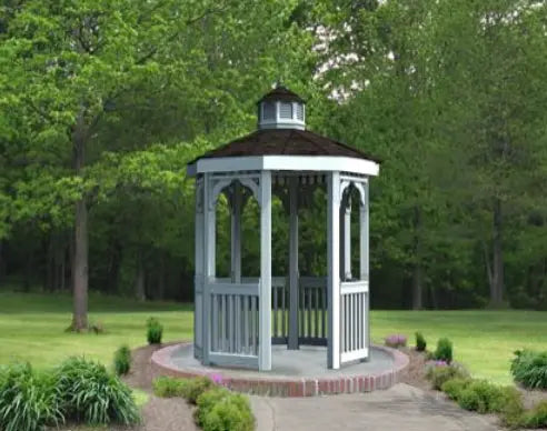 A gray octagonal gazebo with a classic design stands on a brick patio surrounded by greenery. The gazebo features a shingled roof with a cupola, decorative railings, and built-in benches. Trees and shrubs frame the gazebo in the background.