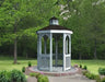 A gray octagonal gazebo with a classic design stands on a brick patio surrounded by greenery. The gazebo features a shingled roof with a cupola, decorative railings, and built-in benches. Trees and shrubs frame the gazebo in the background.