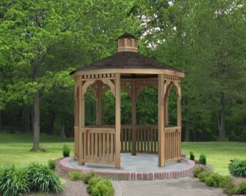 A wooden octagonal gazebo with a shingled roof, decorative cupola, and latticework detailing on the walls and roofline, surrounded by greenery and a brick patio.