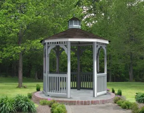 A gray vinyl octagonal gazebo with a classic design stands on a brick patio surrounded by greenery. The gazebo features a shingled roof with a cupola, decorative railings, and built-in benches. Trees and shrubs frame the gazebo in the background.