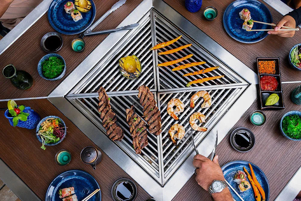 Grill with skewered food on a wooden korean bbq grill table with plates and bowls of ingredients.