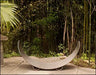 An empty carbon steel crescent log rack placed in a garden setting with bamboo plants and a palm tree in the background.